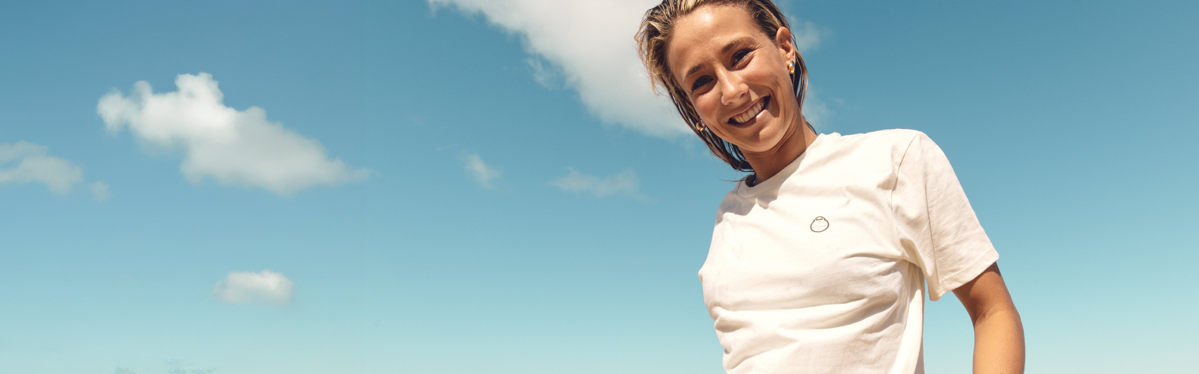 Woman smiling on the beach in a white logo tee under sunny skies, featuring the SB Apparel Collection – Sun Bum.
