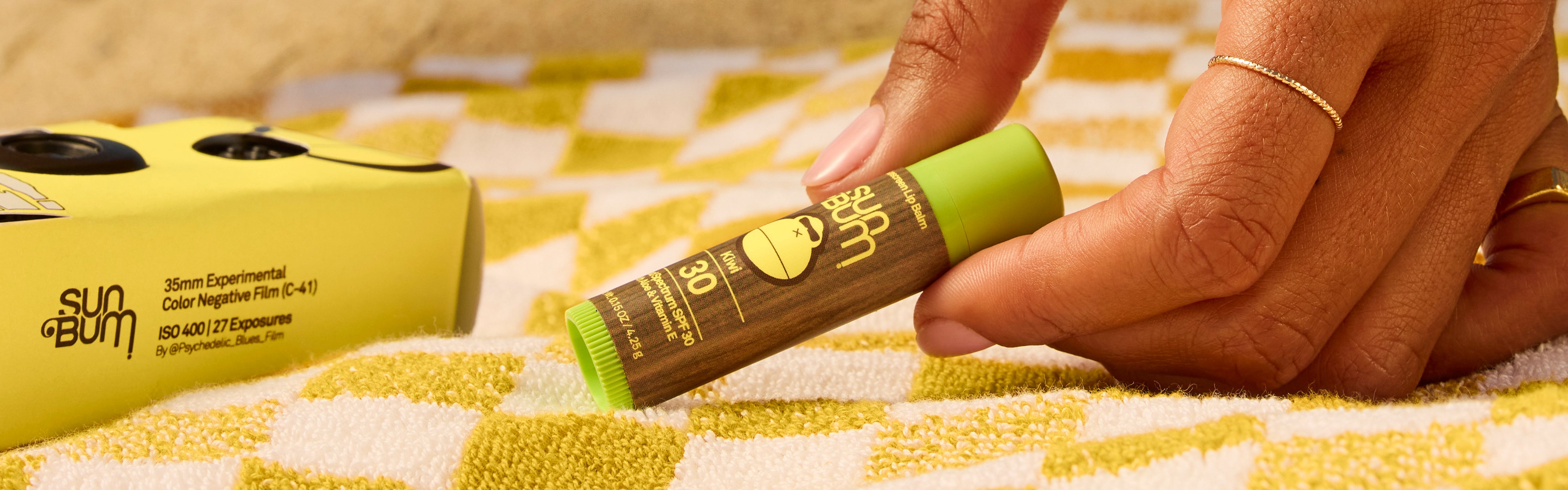 Close-up of hand picking up SPF 30 Kiwi lip balm on a beach towel, from the SB OG Lip Care Collection – Sun Bum.
