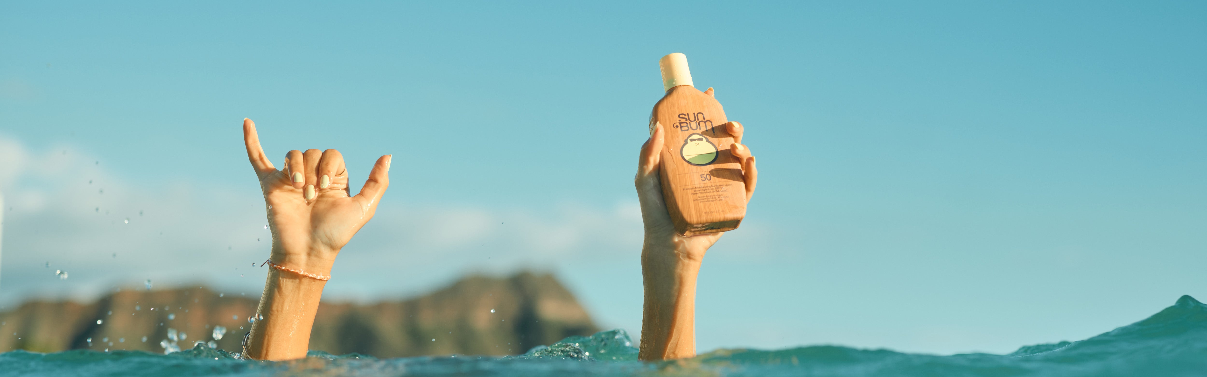 Hands raised from the ocean, one holding SPF 50 lotion, showcasing the Sunscreen Collection – Sun Bum.