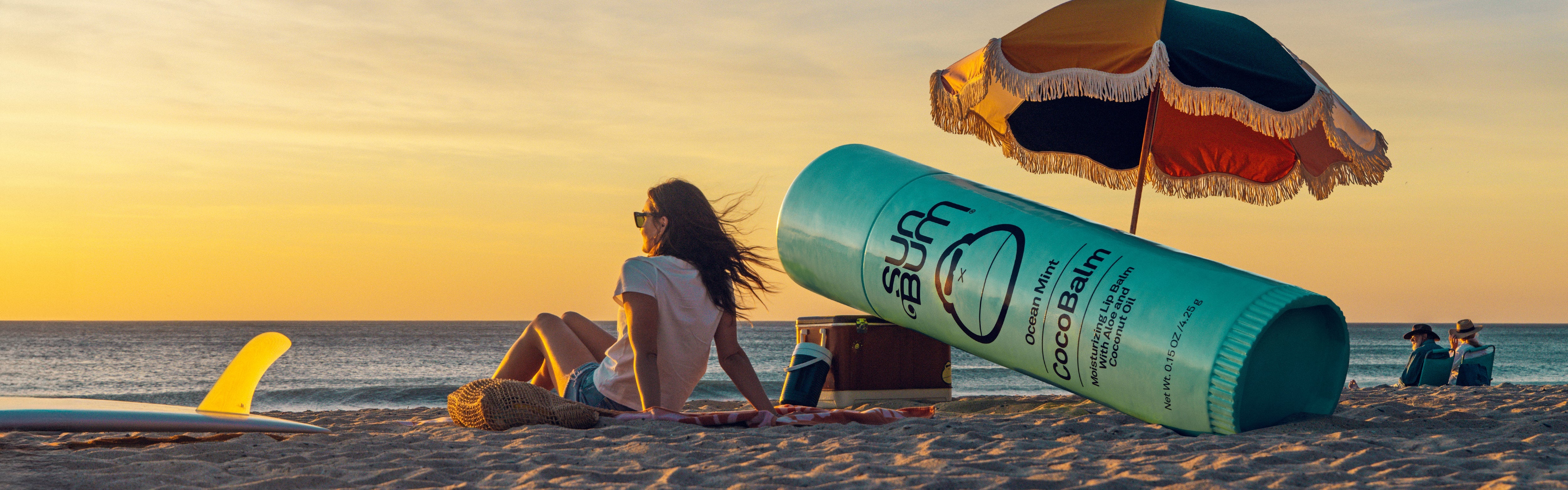 Woman relaxing on the beach at sunset next to oversized CocoBalm lip balm, from the SB CocoBalm Collection – Sun Bum.