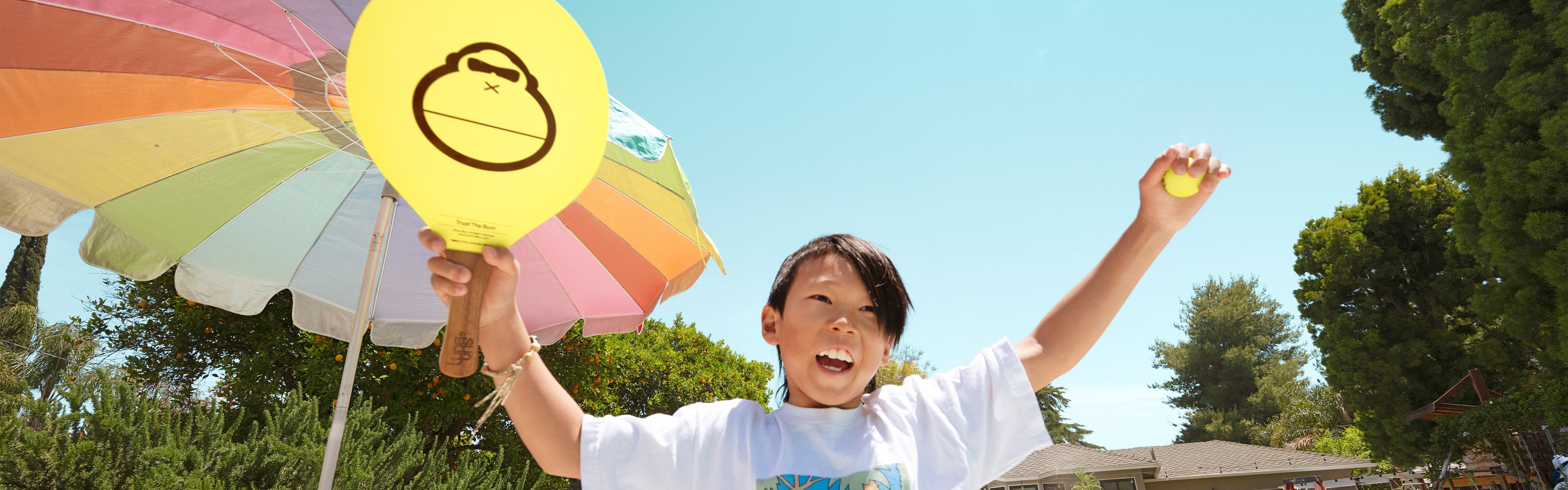 Smiling kid playing paddle ball in Sun Bum gear under a beach umbrella, featuring the SB Merch Collection – Sun Bum.