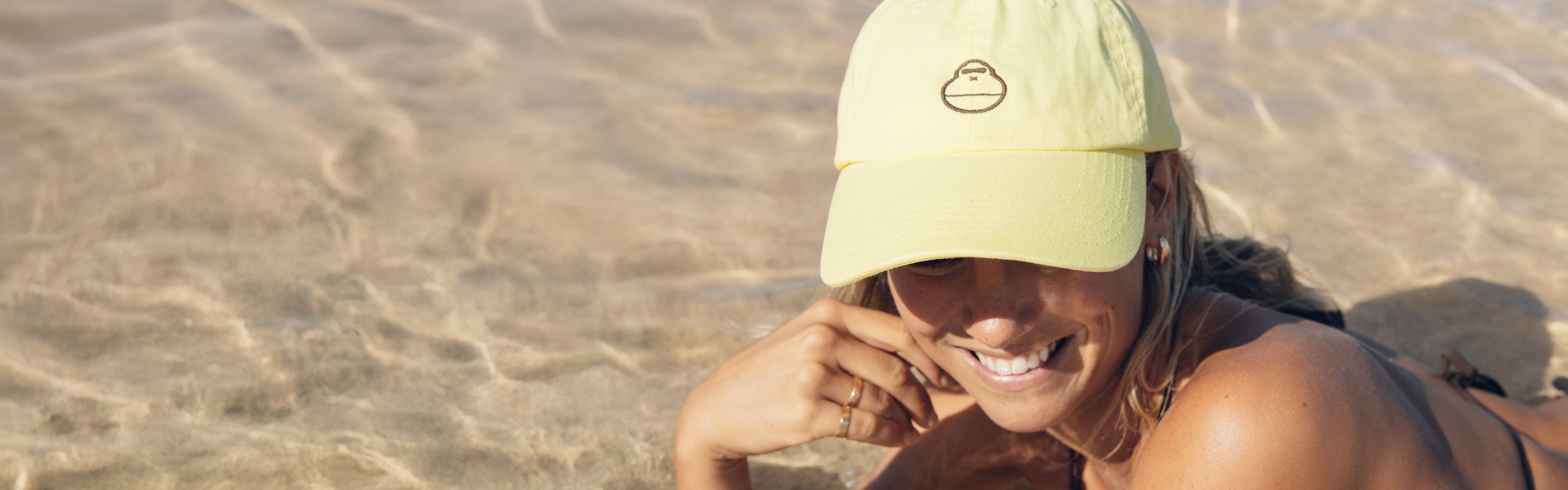 Woman lounging in shallow water wearing a yellow logo cap, featuring the SB Accessories Collection – Sun Bum.