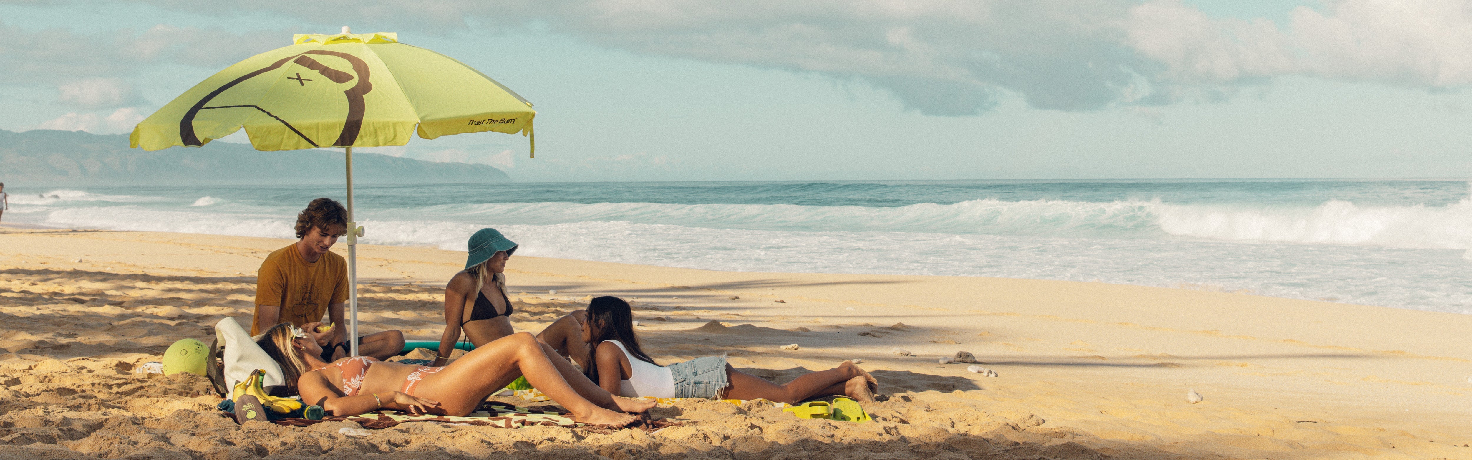 Group of friends relaxing under a Sun Bum umbrella on the sand, featuring the SB Beach Day Essentials Collection – Sun Bum.