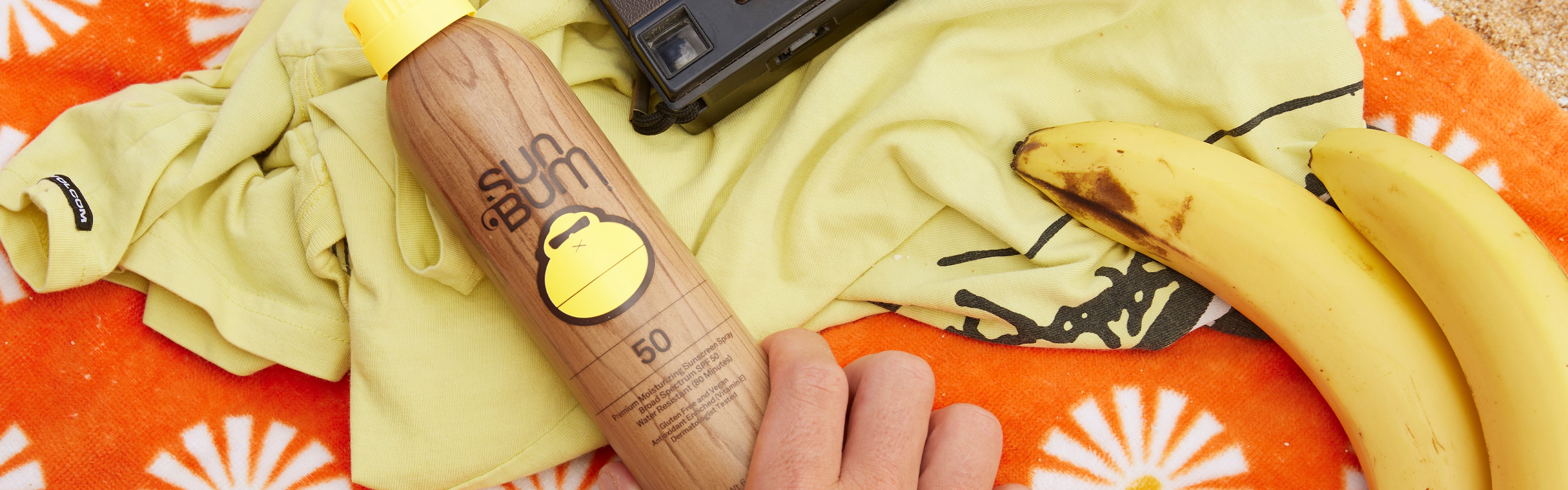 Hand holding Sun Bum SPF 50 spray next to vintage camera on beach towel – Sunscreen Sprays and Mist Collection - Sun Bum.