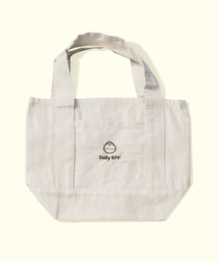 A light beige canvas tote bag with dual shoulder straps and a front pocket, featuring a small black embroidered logo that reads 'Daily SPF' with a droplet icon, set against a solid cream background.
