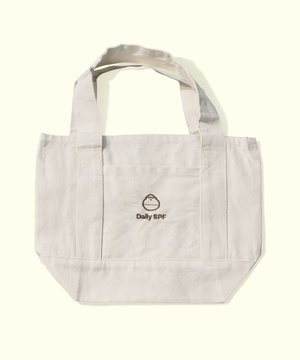 A light beige canvas tote bag with dual shoulder straps and a front pocket, featuring a small black embroidered logo that reads 'Daily SPF' with a droplet icon, set against a solid cream background.