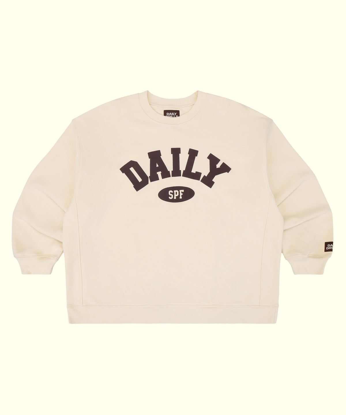 An off-white crewneck sweatshirt featuring a dark brown graphic on the chest that reads 'DAILY SPF' with 'SPF' inside an oval. A small brand tag is visible on the left sleeve cuff, and the inner collar shows the brand name 'DAILY'.