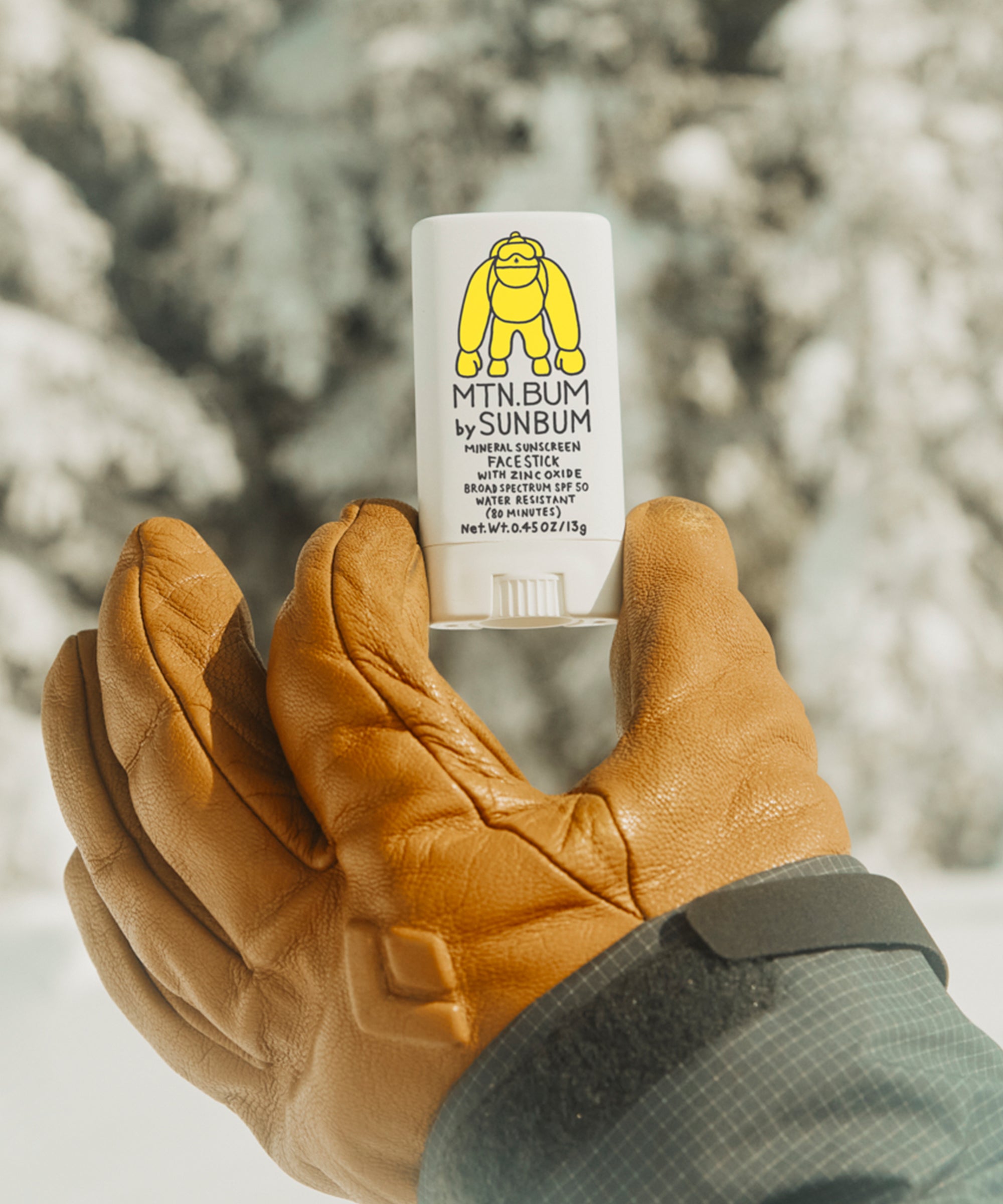 Hand wearing a brown glove holding a white container labeled 'MTN BUM by SUNBUM' against a snowy background.