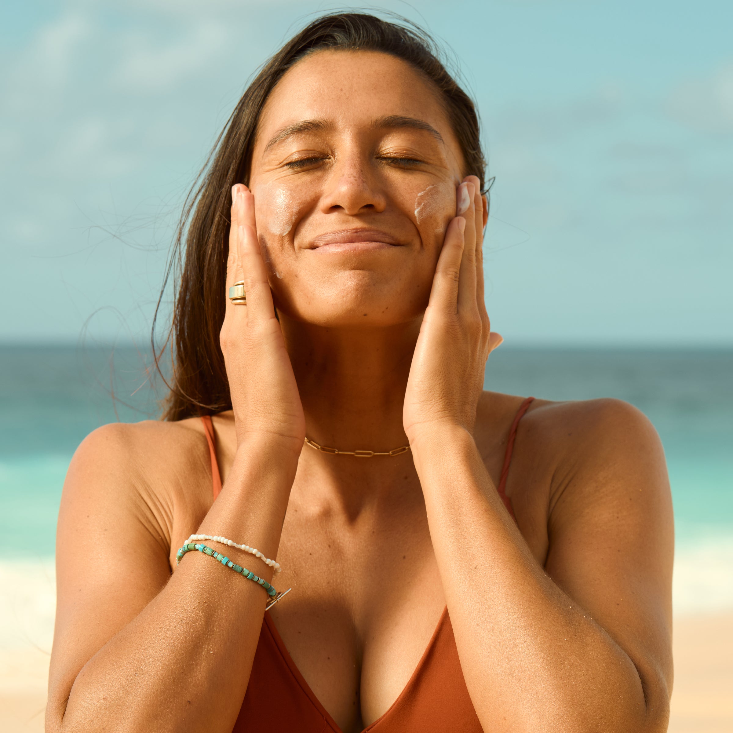 Woman applying Sun Bum face sunscreen by the ocean, promoting broad spectrum daily sun protection - Original SPF Collection - Sun Bum