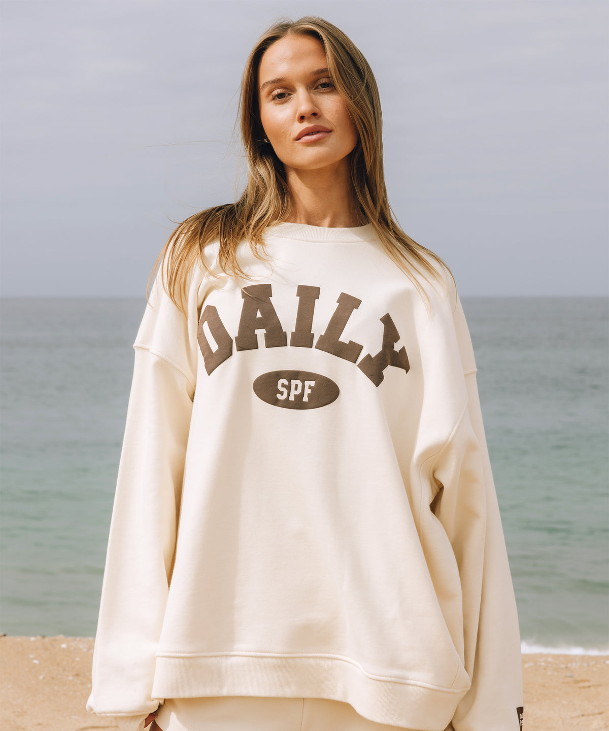 A woman standing on a beach wearing an oversized cream sweatshirt with 'DAILY SPF' printed in brown across the front.