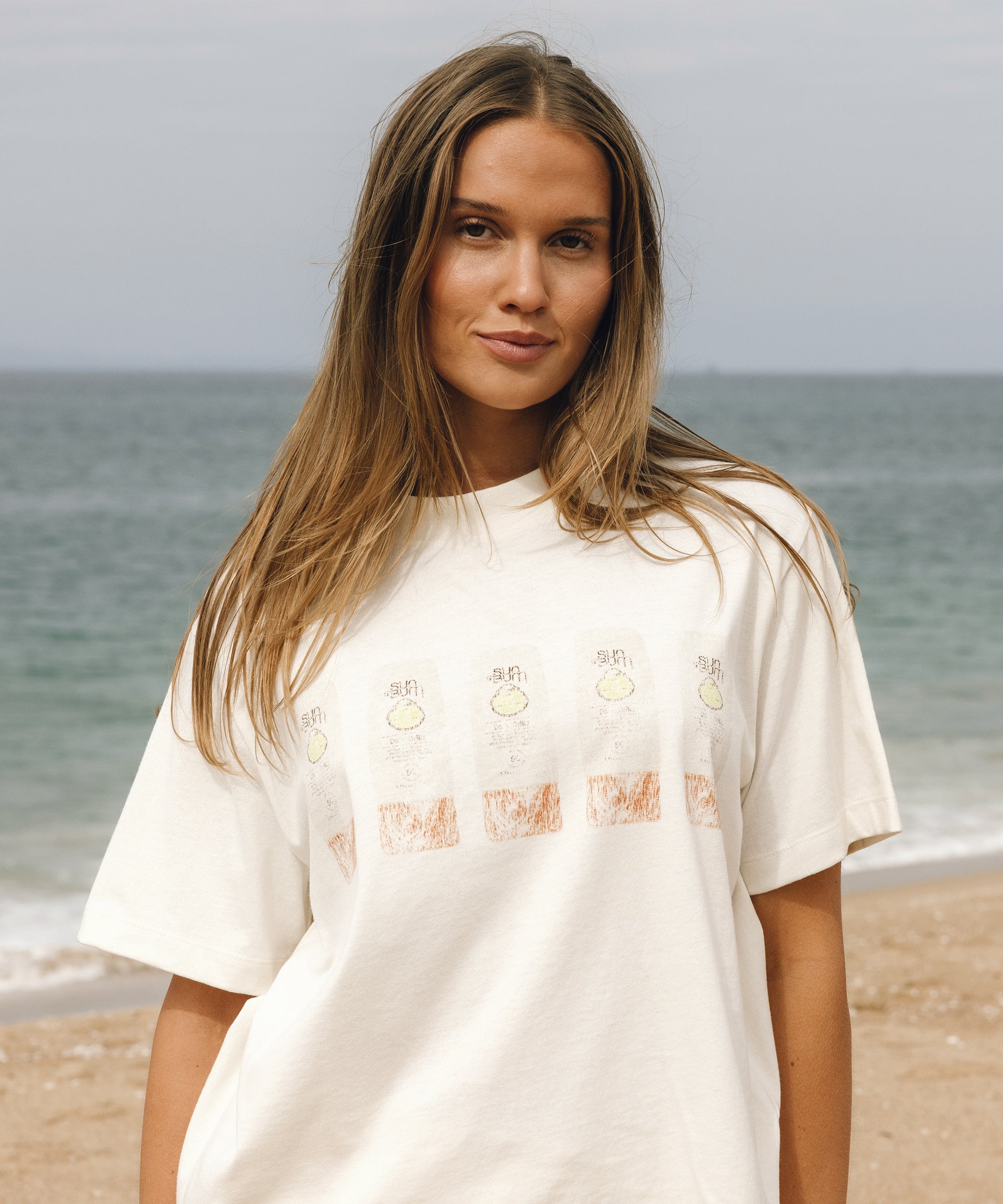A woman with long light brown hair stands on a sandy beach wearing an oversized off-white t-shirt with a graphic print of lemons and orange squares across the chest.