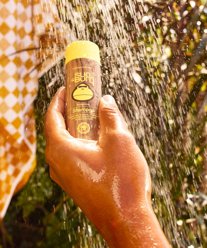 Revitalizing Shampoo bottle held up against sparkling water droplets in sunlight, showcasing the wooden-toned packaging with yellow cap and monkey logo. Natural outdoor setting with bokeh effects and warm golden tones. By Sun Bum.