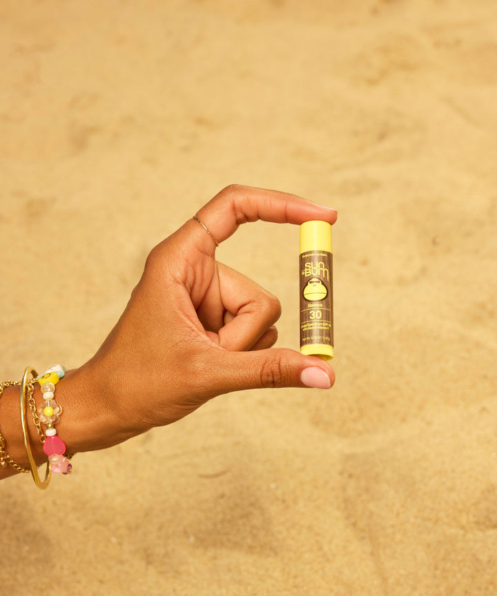 Hand holding Original SPF 30 Sunscreen Lip Balm in Banana flavor against sandy beach background, featuring colorful beaded bracelets. Product shows moisturizing lip protection for sun-safe summer days by Sun Bum.