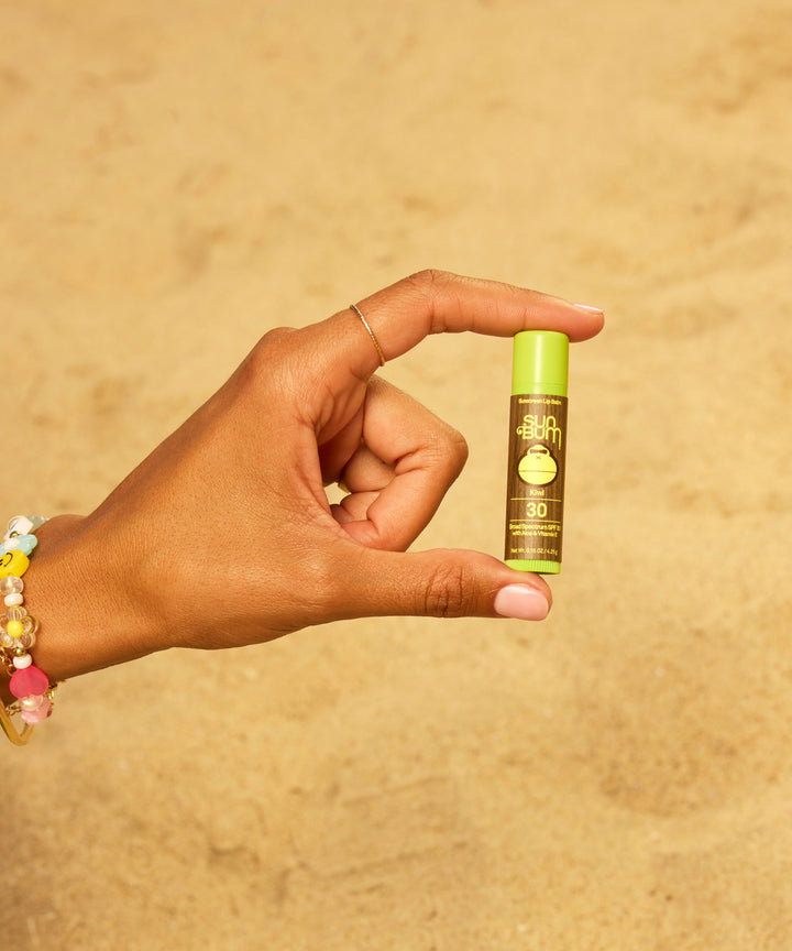 Hand holding Original SPF 30 Sunscreen Lip Balm in Kiwi flavor against a warm sandy background, featuring bright green packaging and colorful beaded bracelet accessory by Sun Bum