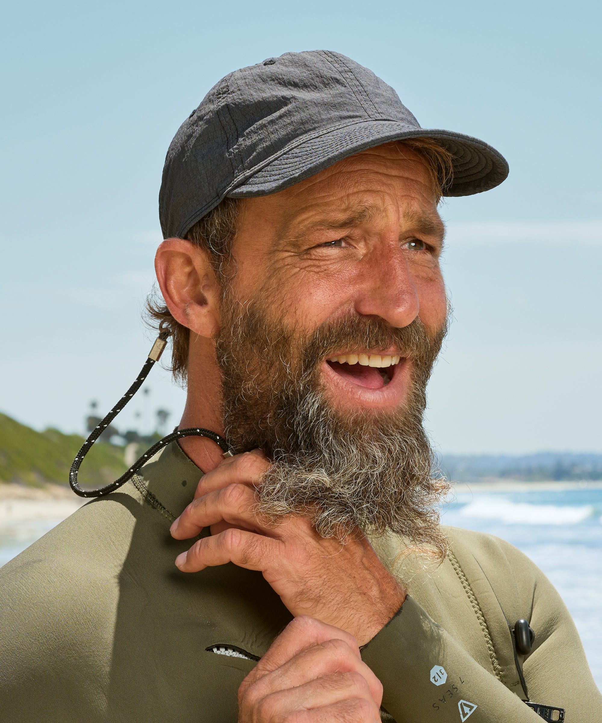 Man with a beard and cap standing by the water, wearing a wetsuit.