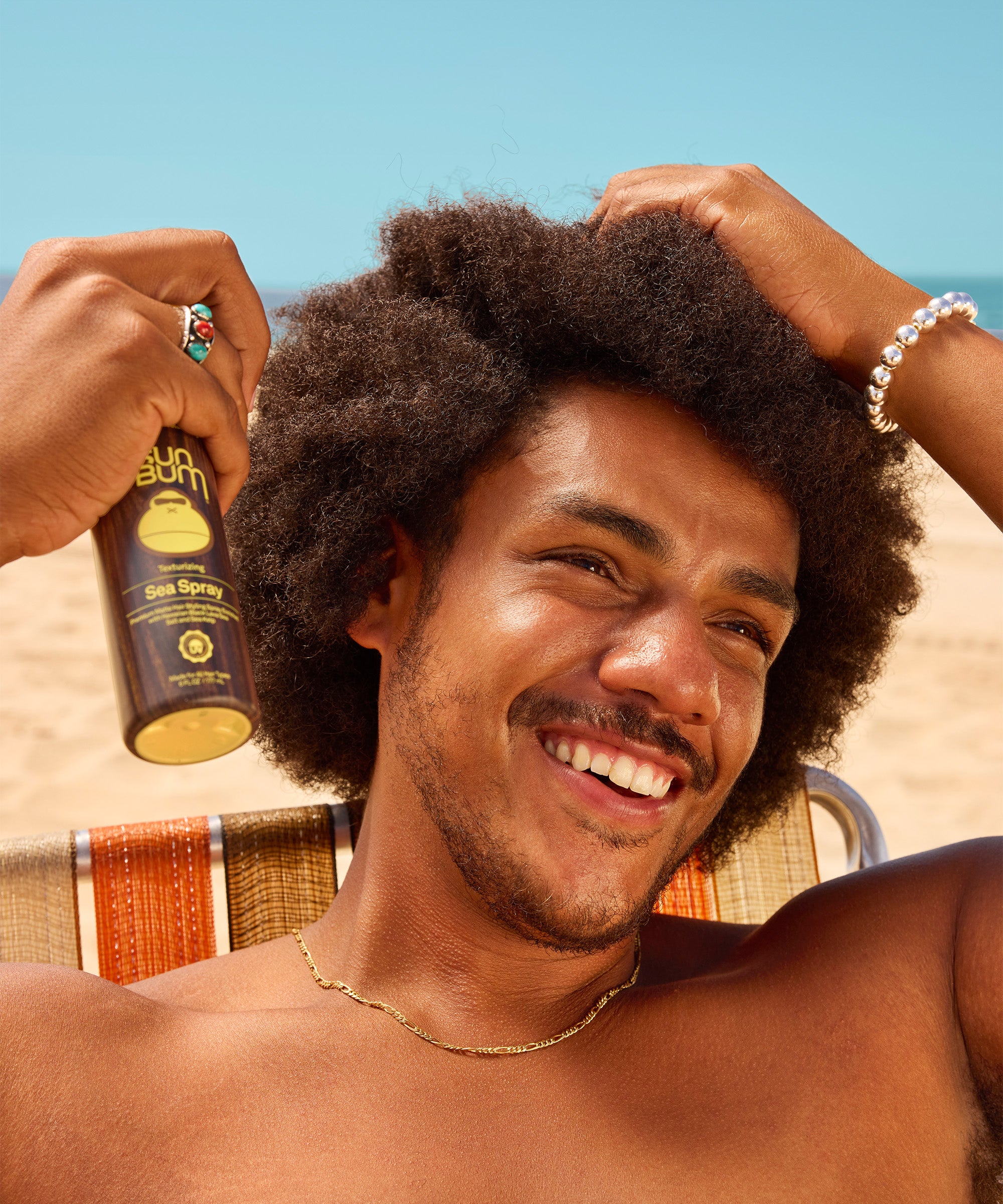 Sea Spray texturizing hair product shown on a bright sunny day, featuring a brown bottle with yellow accents against a blue sky. Perfect for achieving natural beach-styled hair texture by Sun Bum.