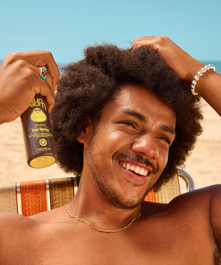 Sea Spray texturizing hair product shown on a bright sunny day, featuring a brown bottle with yellow accents against a blue sky. Perfect for achieving natural beach-styled hair texture by Sun Bum.