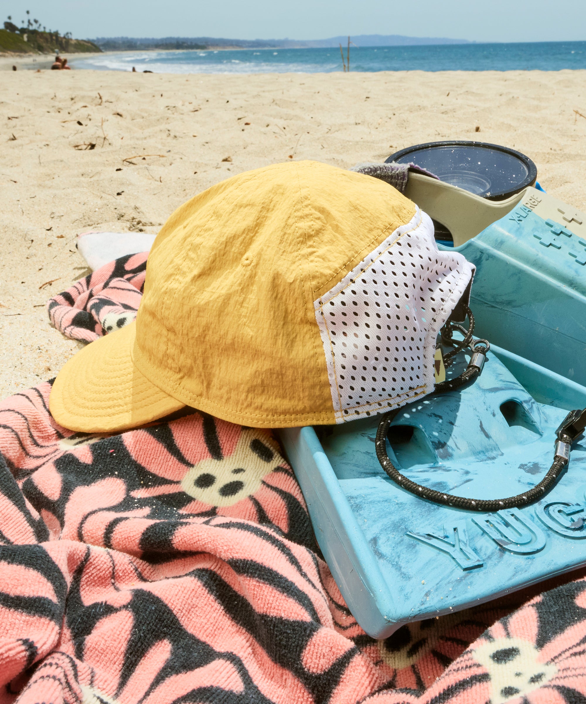 Yellow cap, sunglasses, and pink towel on a beach with a blue cooler.