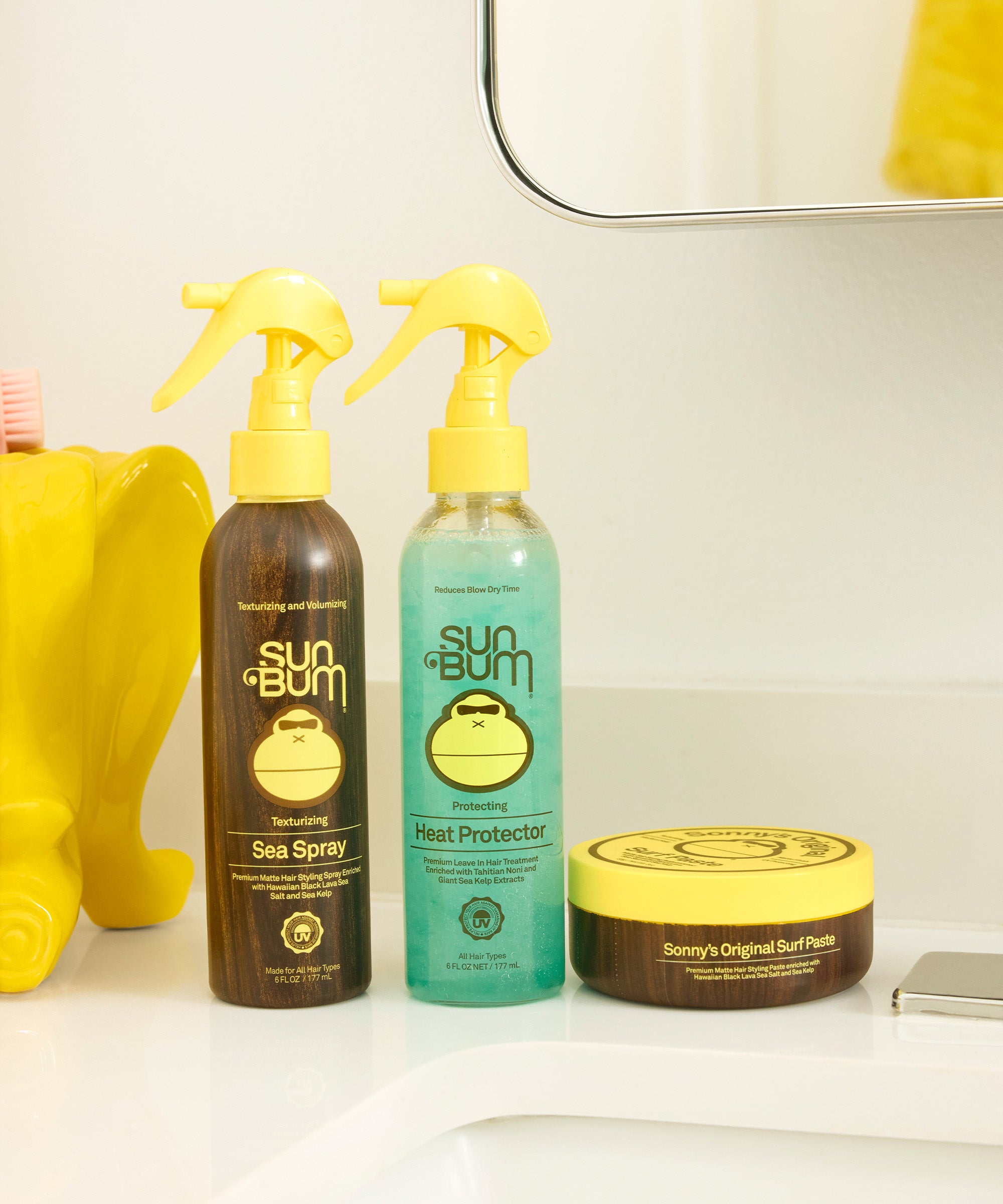 Heat Protector spray bottle in turquoise blue alongside brown Sea Spray and hair paste containers, featuring Sun Bum's signature monkey logo on a clean white bathroom counter. By Sun Bum.