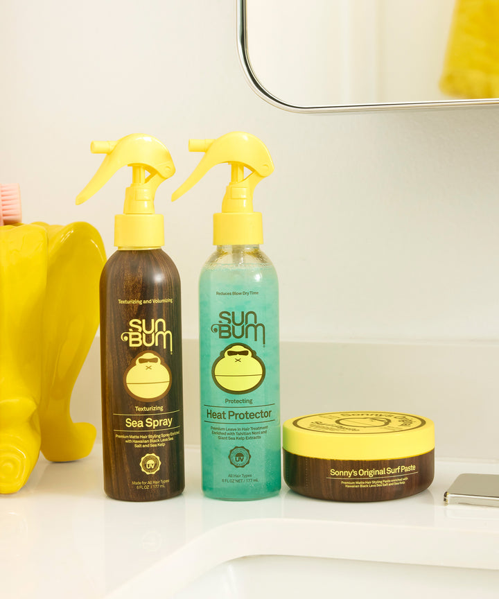 Heat Protector spray bottle in turquoise blue alongside brown Sea Spray and hair paste containers, featuring Sun Bum's signature monkey logo on a clean white bathroom counter. By Sun Bum.