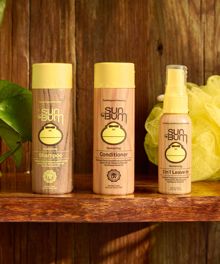 Three Sun Bum hair care products, including Revitalizing Shampoo, displayed on wooden shelf with yellow loofah and green leaf, featuring clean pink and yellow packaging with monkey logo - by Sun Bum