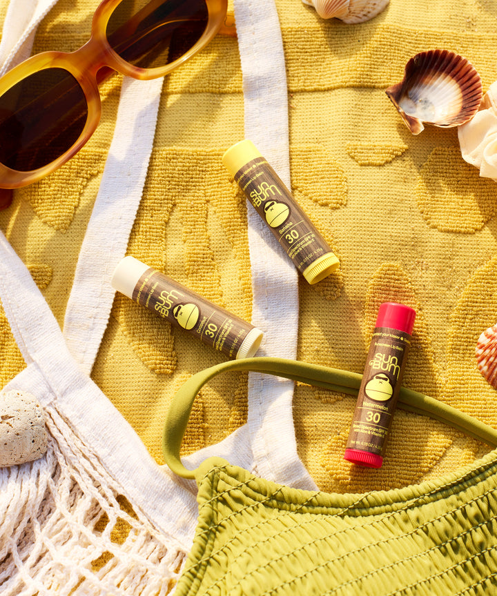 Three Original SPF 30 Sunscreen Lip Balms displayed on a yellow beach towel with sunglasses, seashells, and a green swimsuit top, creating a summer beach-ready scene featuring Sun Bum's protective lip care essentials.