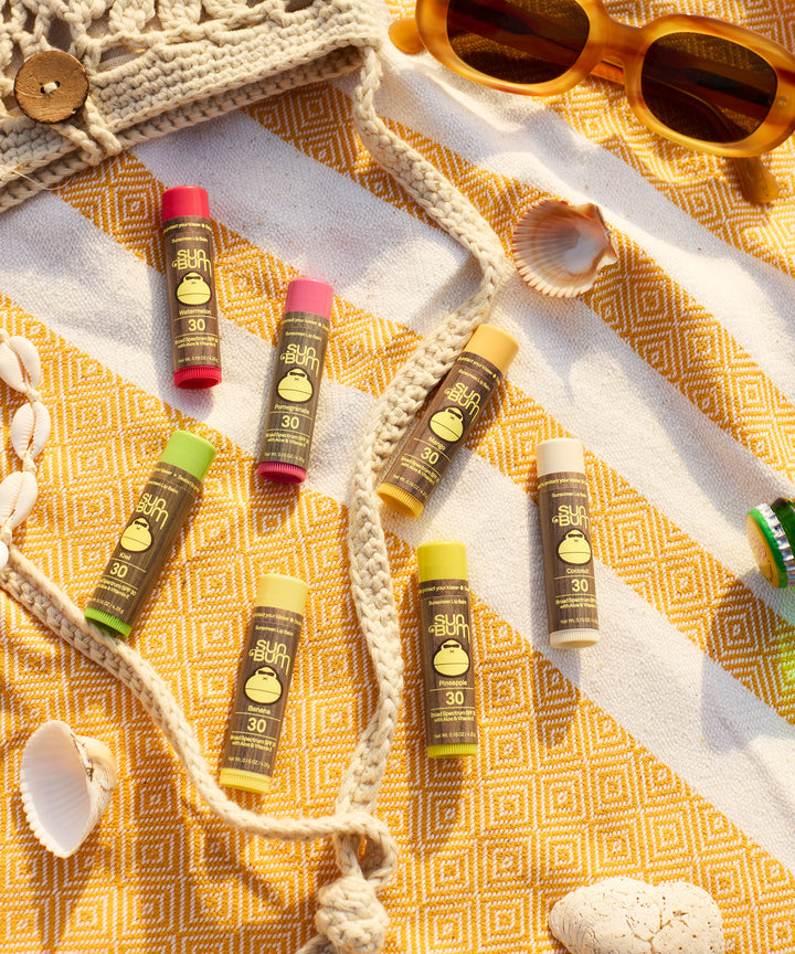 Original SPF 30 Sunscreen Lip Balms displayed on a yellow geometric patterned beach blanket with seashells and sunglasses, showcasing various flavors including banana in colorful tubes by Sun Bum