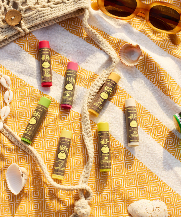 Original SPF 30 Sunscreen Lip Balm in Kiwi flavor displayed on a yellow patterned beach blanket with seashells, rope, and yellow sunglasses, showcasing Sun Bum's colorful lip care collection for summer protection - by Sun Bum