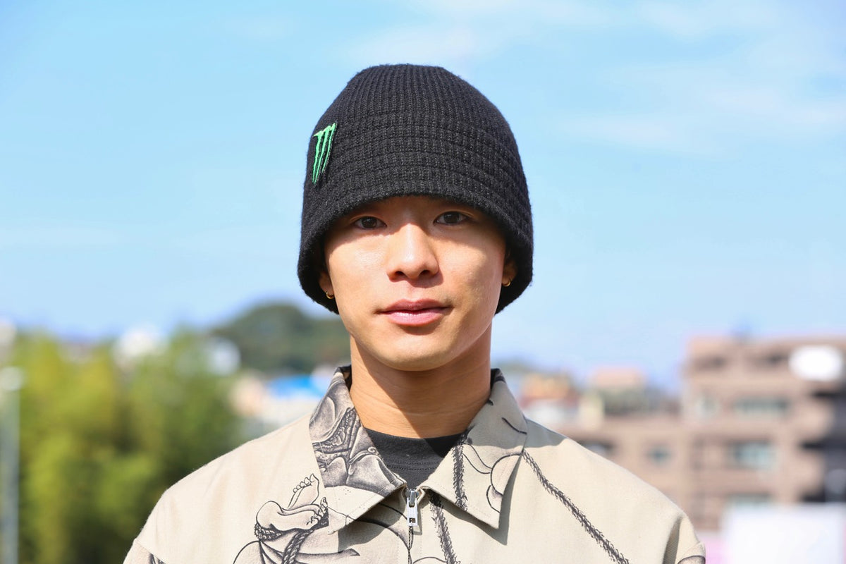 A man wearing a black knit beanie with a green Monster Energy logo and a beige patterned jacket, photographed outdoors against a blurred blue sky and buildings.