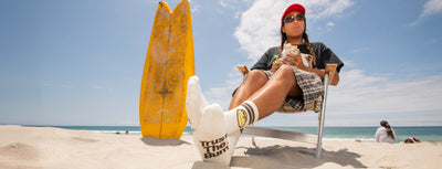 Person sitting on a beach with a surfboard, wearing sunglasses and a hat.