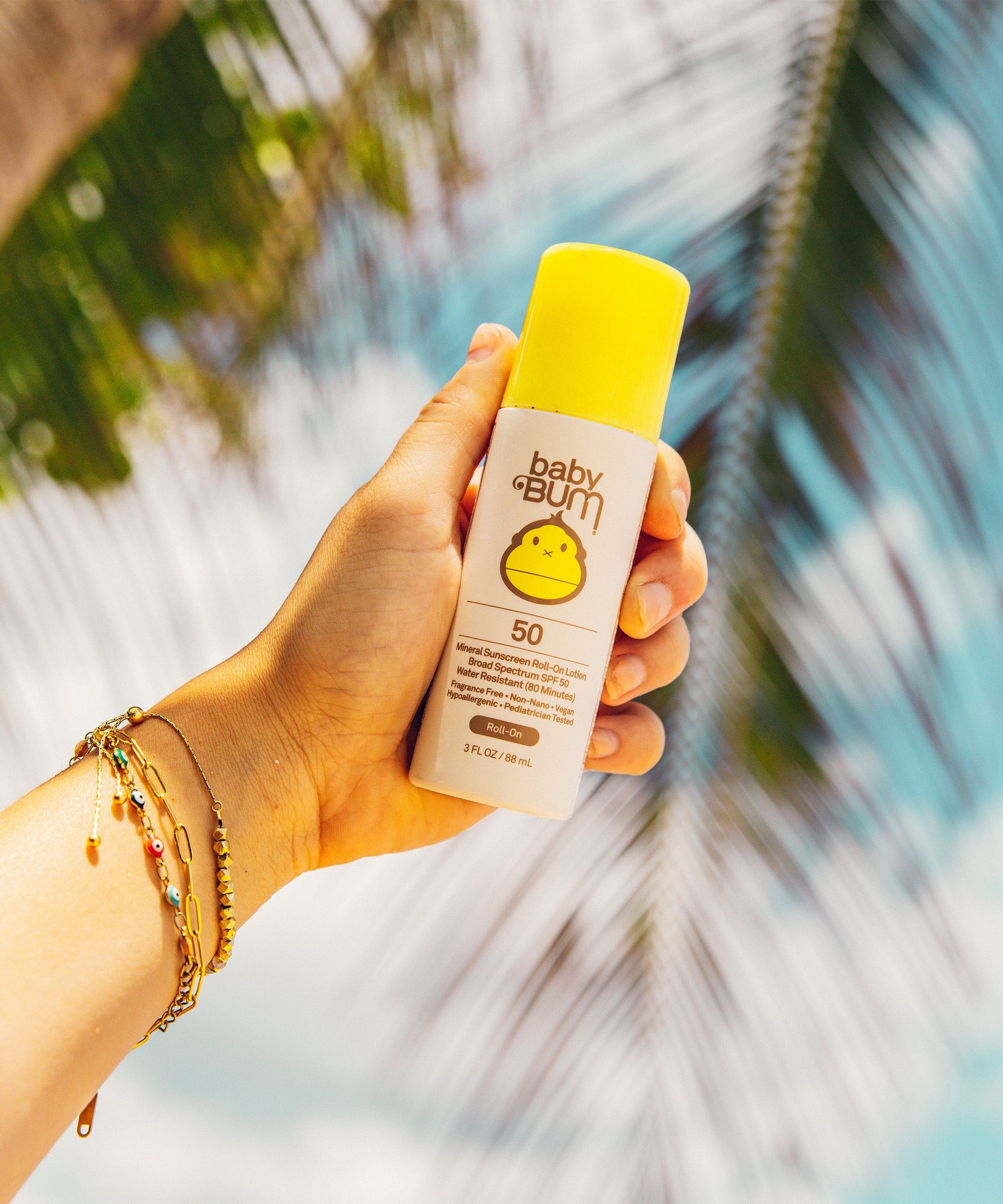 Hand holding Baby Bum Mineral SPF 50 Roll-On Sunscreen with palm tree background by Baby Bum