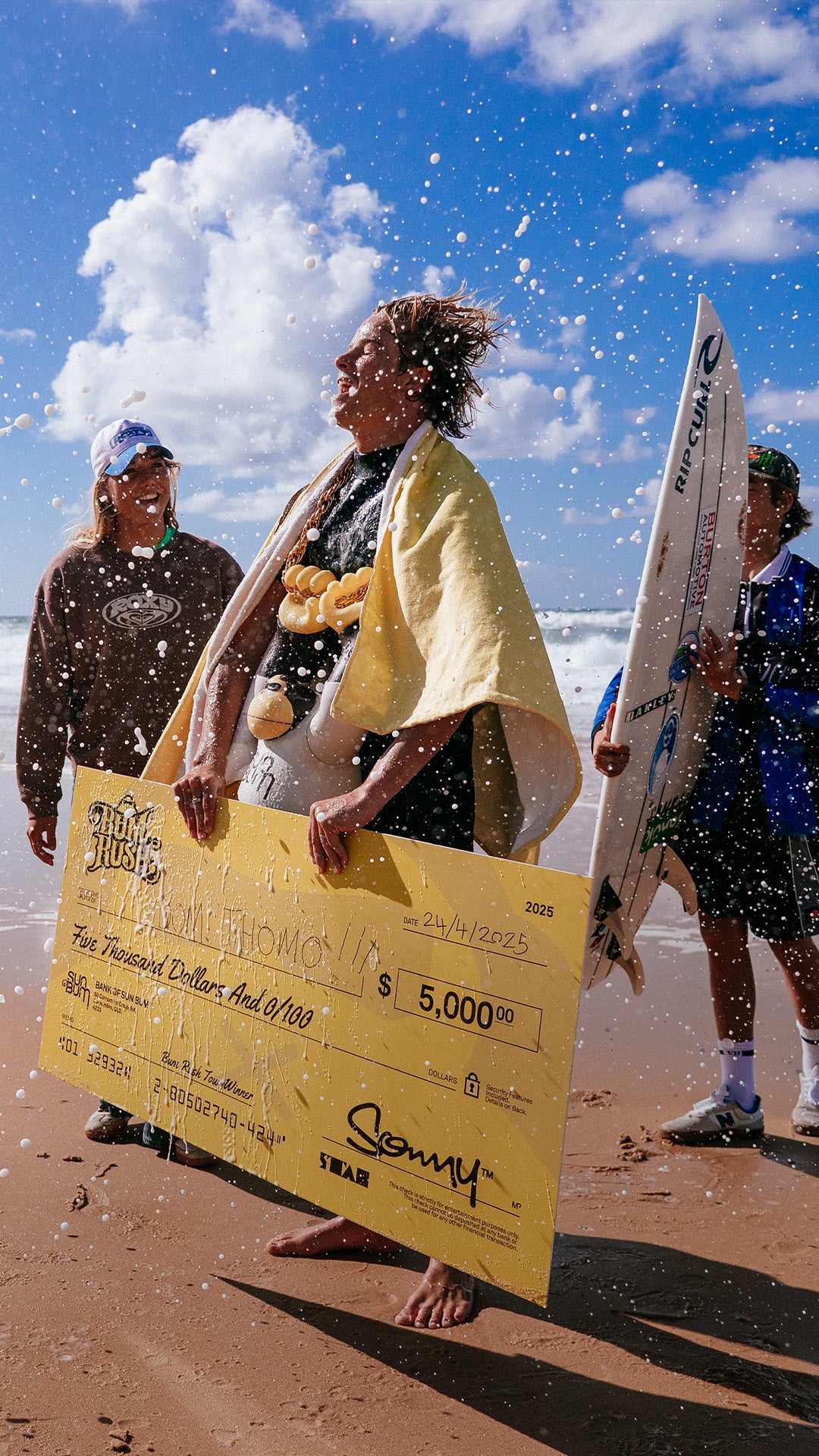 Person holding a large check on a beach with surfboards and people in the background