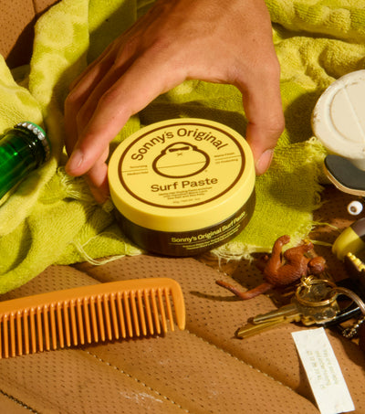 Hand holding a container of Sonny's Original Surf Paste on a textured surface with a comb and keys.