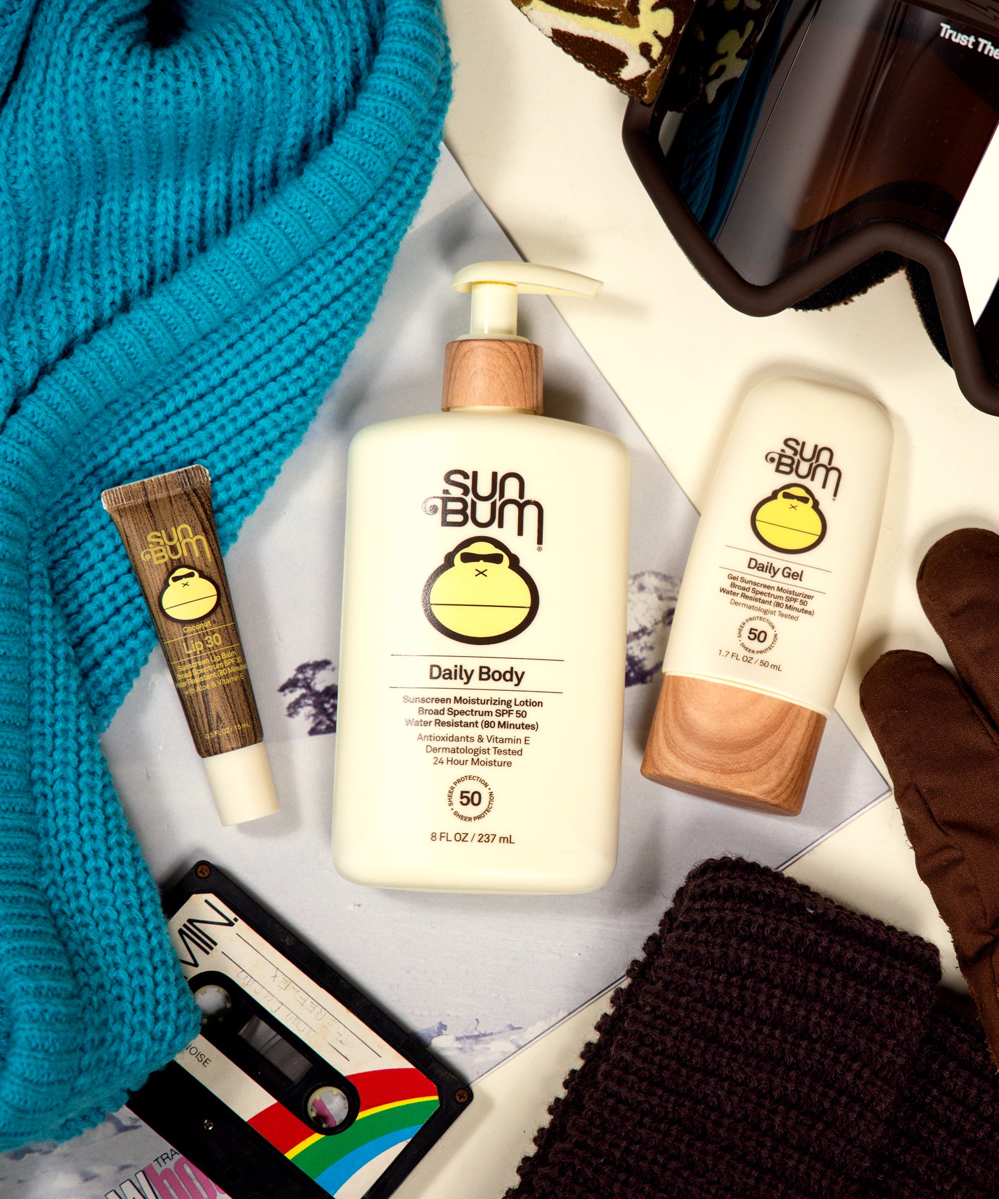 Sun Bum sunscreen products with gloves, scarf, and sunglasses on a textured surface