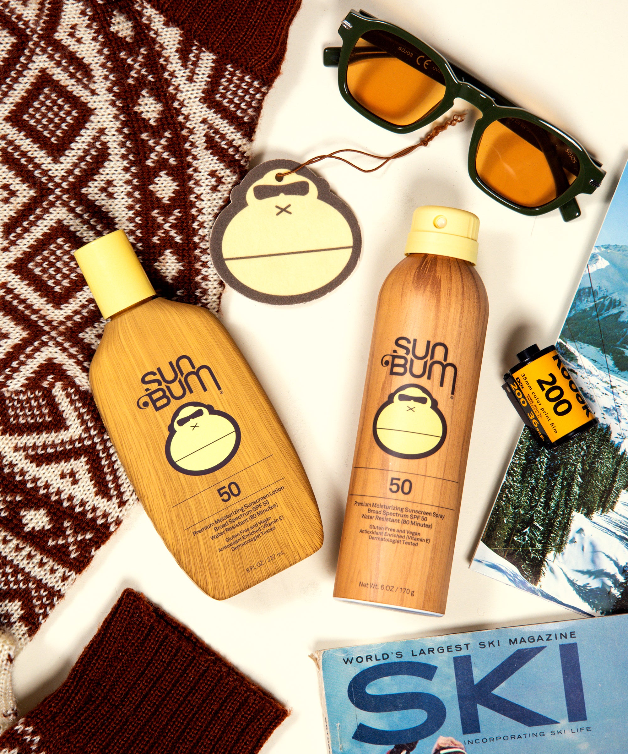 Two bottles of Sun Bum sunscreen, a pair of sunglasses, and a magazine titled 'World's Largest Ski Magazine' on a patterned surface.
