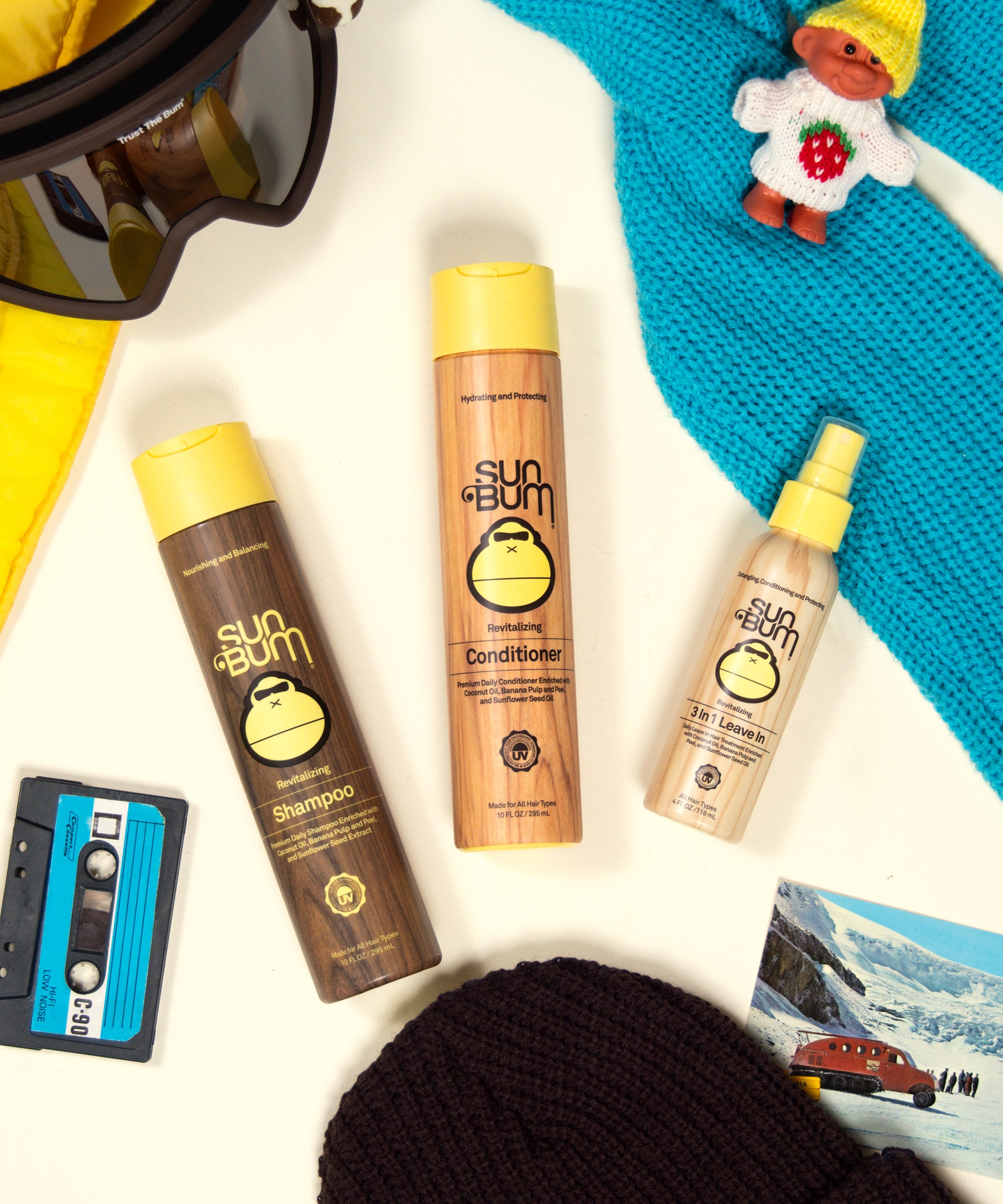 Sun Bum shampoo, conditioner, and spray bottles on a white surface with a blue towel and rubber duck toy.