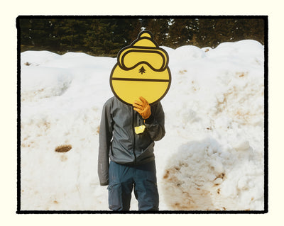 Person wearing a yellow mask with goggles and a tree design, standing in a snowy landscape.