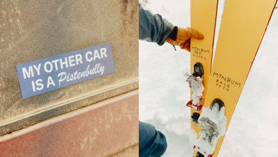 Car with a sticker 'My other car is a Pistenbully' and a person holding yellow skis in the snow.
