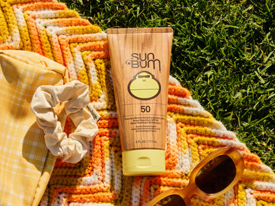 Sun Bum sunscreen tube on a colorful towel with sunglasses and a scrunchie in the background.