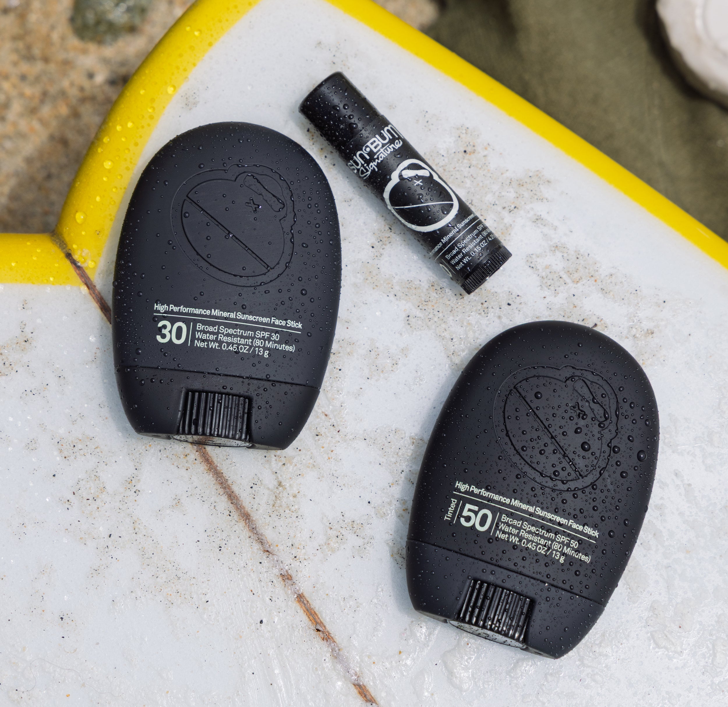 Two black Sun Bum Signature sunscreens on a textured surf board with a blurred beach background