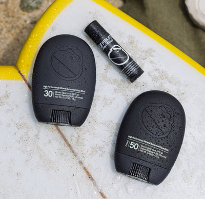 Two black Sun Bum Signature sunscreens on a textured surf board with a blurred beach background