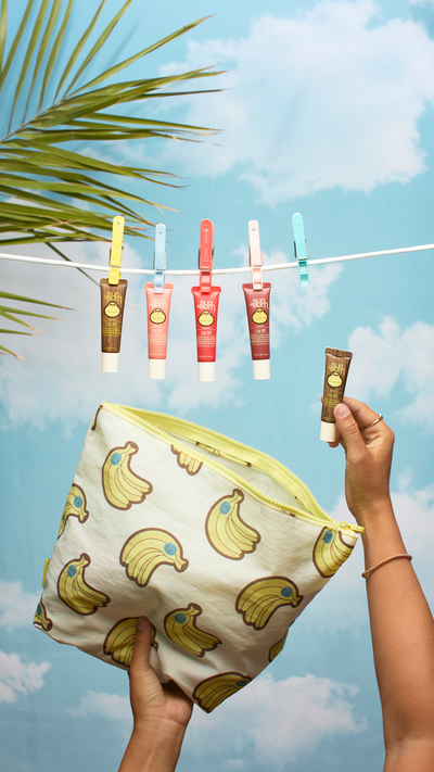 A person holds an open cream-colored zip pouch with a yellow zipper and a banana print, while another hand holds a small brown tube. Five similar cosmetic tubes hang from a white clothesline against a blue sky and clouds backdrop with a palm leaf in the corner.
