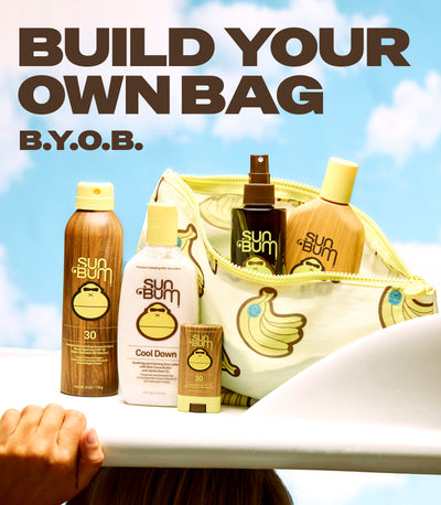 A promotional product shot showing several Sun & Bum branded personal care items arranged beside an open zip pouch with a banana print, set against a bright blue sky background. Large text at the top reads 'BUILD YOUR OWN BAG B.