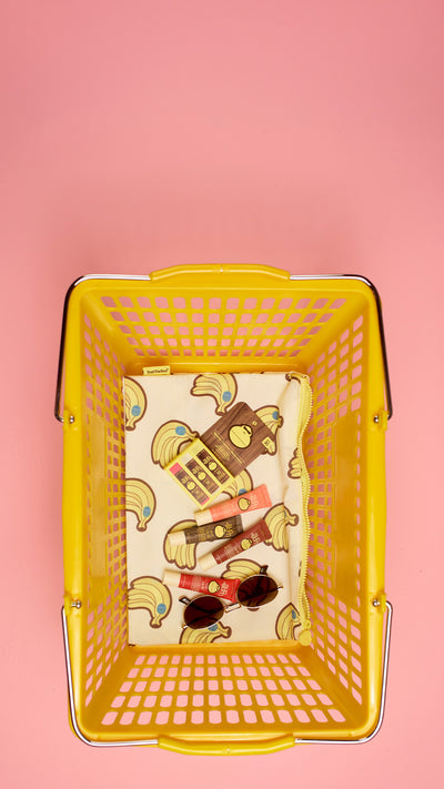 A yellow plastic shopping basket viewed from above, containing a patterned paper insert with illustrations of bananas, a clock, and shopping bags, along with a few small accessories. The basket is set against a solid pink background.