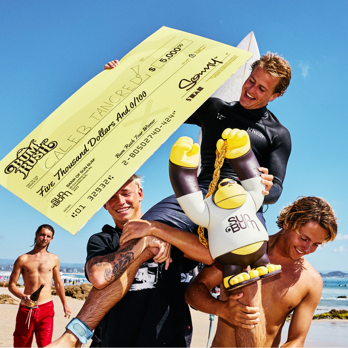 Four men on a beach holding a large yellow check and a cartoon mascot figure. One man wears a black wetsuit, and the background shows blue sky and ocean.
