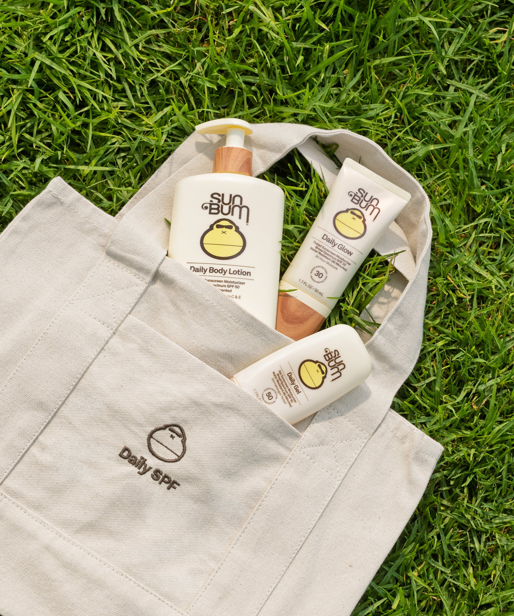 A beige canvas tote bag with 'Daily SPF' printed on the front pocket, filled with sunscreen and body care products from sun bum, placed on a green grass background.