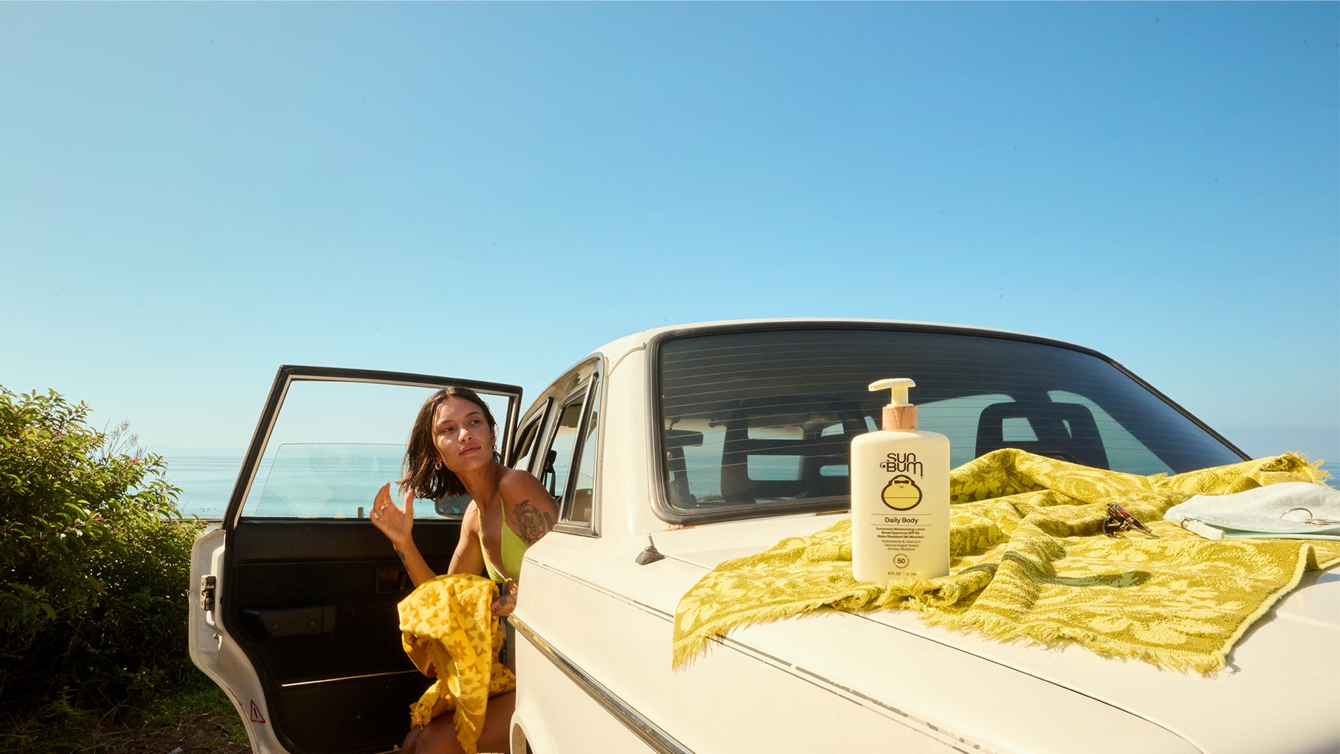 Woman sitting in a car with a bottle of shampoo on a towel, outdoors.