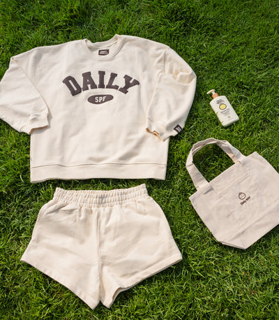 A coordinated outdoor outfit laid flat on green grass, including an off-white sweatshirt with dark gray 'DAILY SPF' text, matching off-white shorts, a small off-white tote bag with a logo, and a small sunscreen bottle.