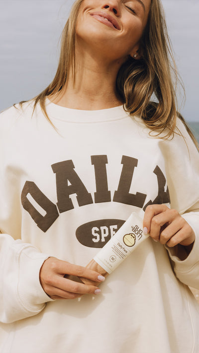 A woman wearing a cream sweatshirt with 'DAILY SPF' printed in dark brown letters, holding a matching SPF tube. The background is a soft, out-of-focus blue-gray sky.