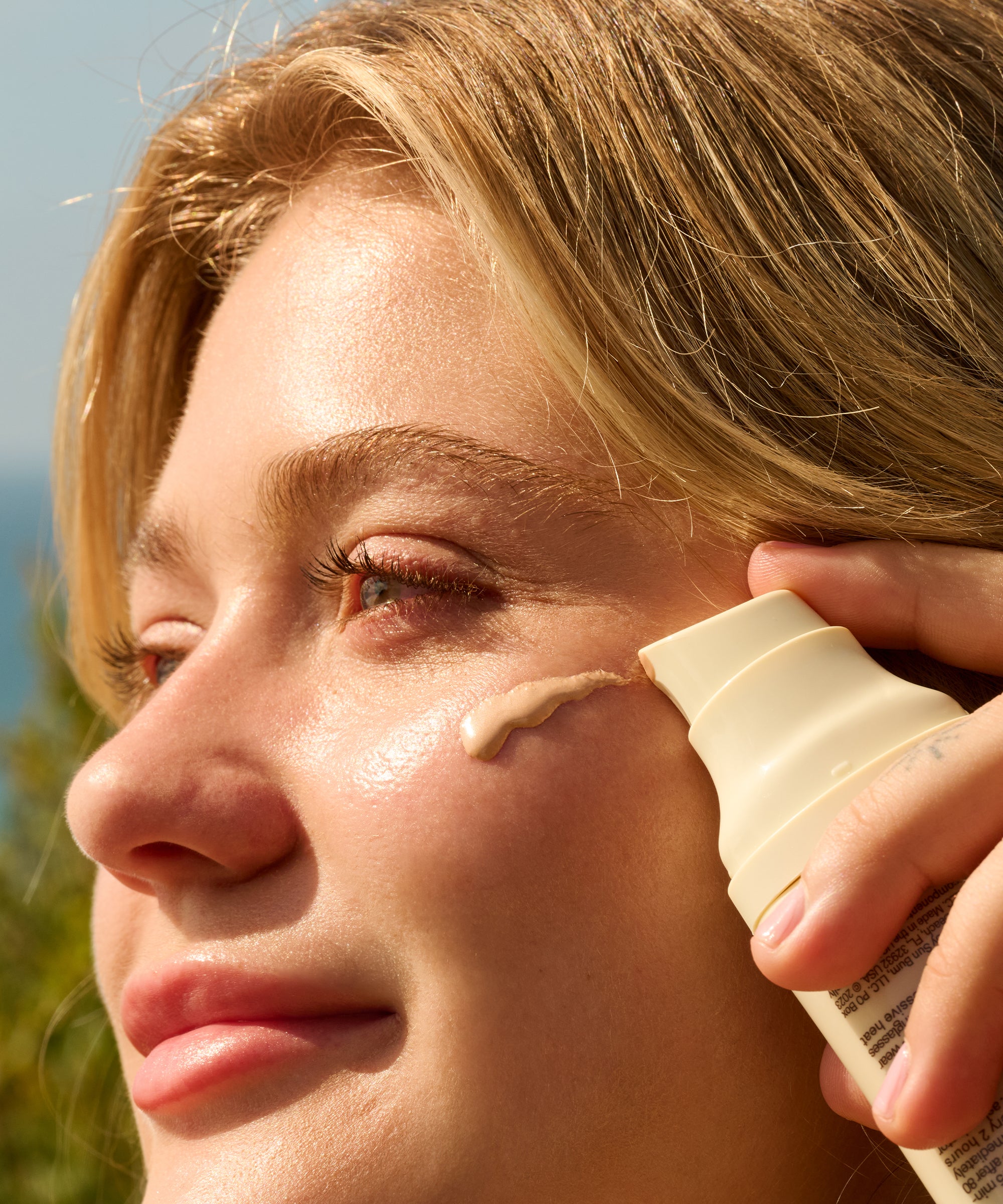 Close-up of a woman applying Daily Body + Glow Bundle cream by Sun Bum on her glowing cheek outdoors.
